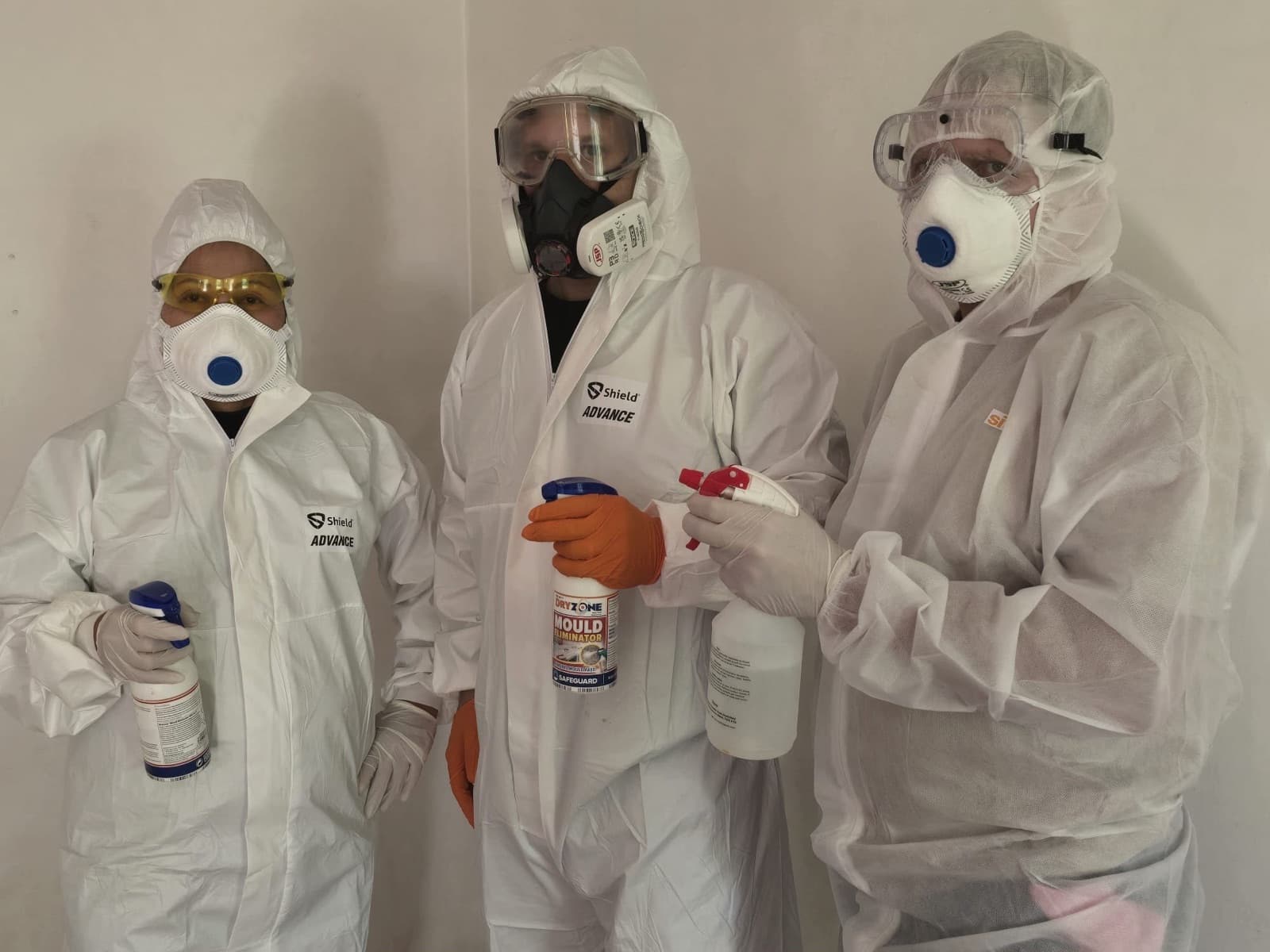Three team members in full protective suits, respirators, goggles and gloves, holding specialist mould treatment products